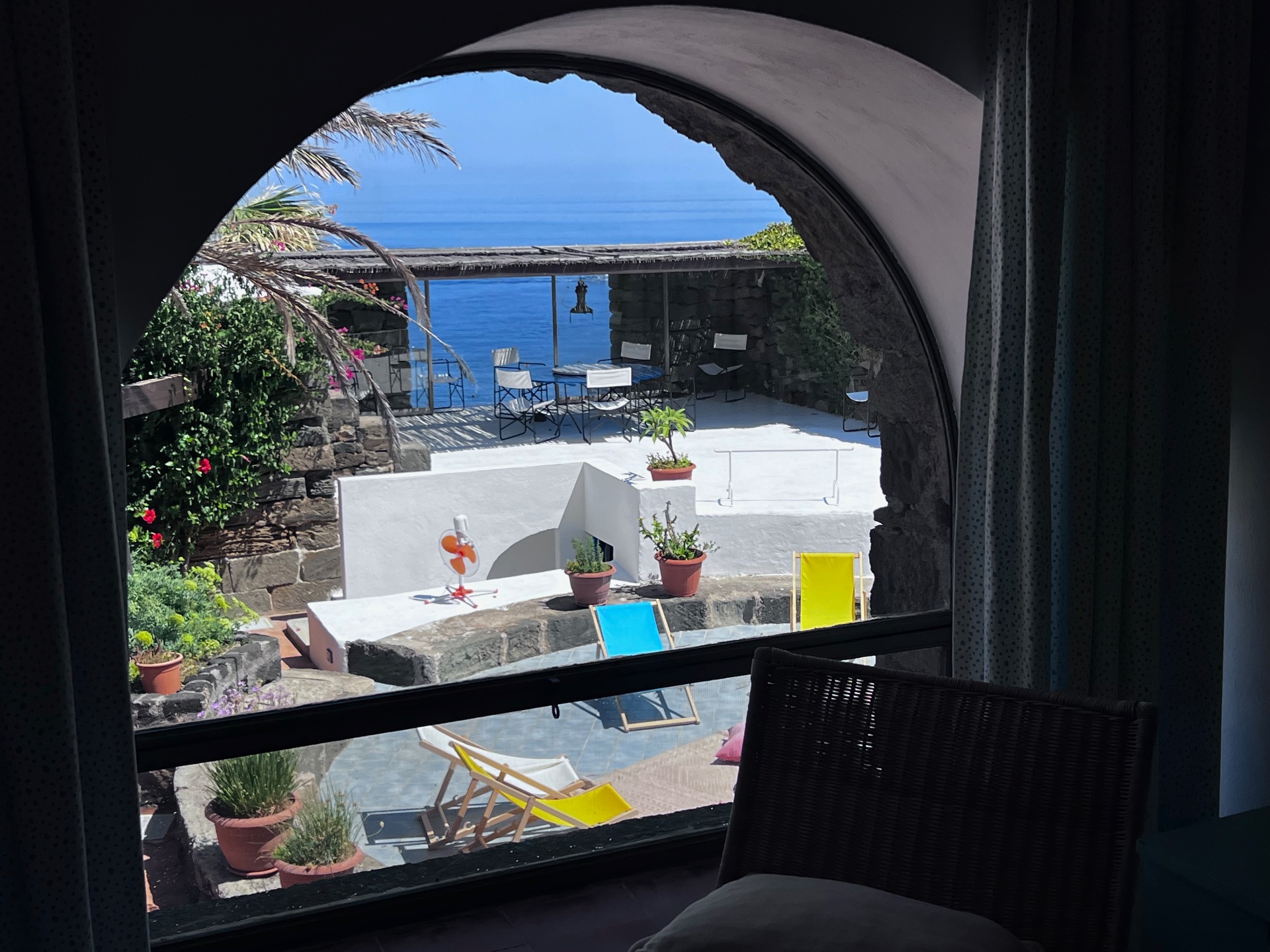View through an arched window of the Royal Suite onto a sunlit terrace with colorful deck chairs and the sea beyond
