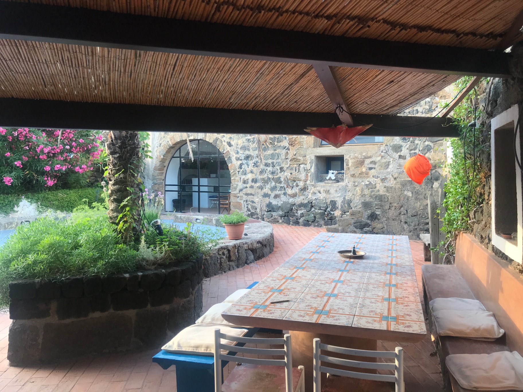 Ceramic-tiled outdoor dining table under bamboo pergola with stone arch and oleander