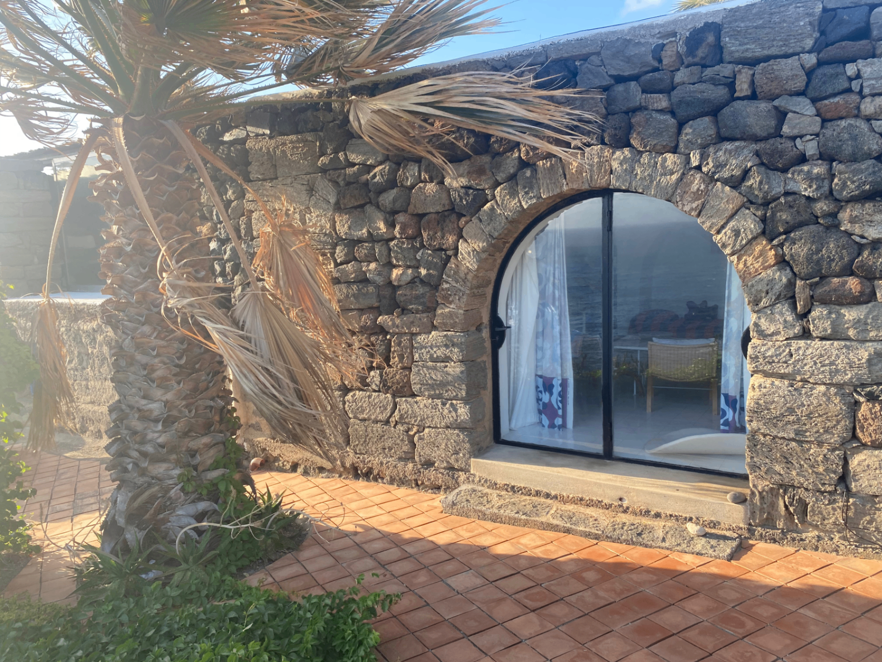 Stonehouse exterior with arched glass door, lava stone walls, palm tree, and terrace