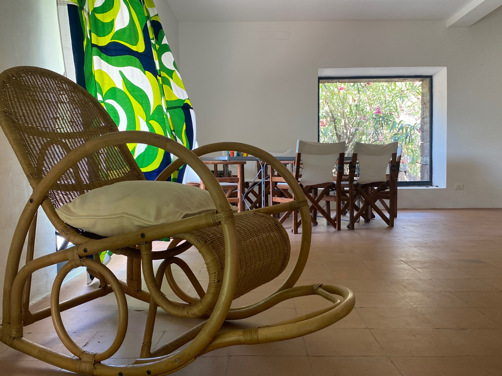 Rattan rocking chair beside a square window framing oleander, with dining table in the background