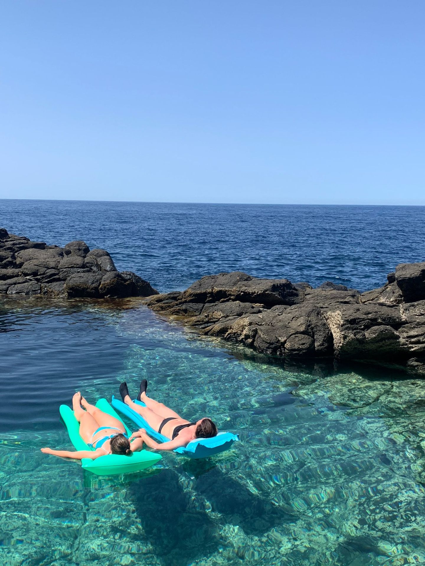 Two women floating on inflatable rafts in a crystal-clear natural rock pool beside the sea