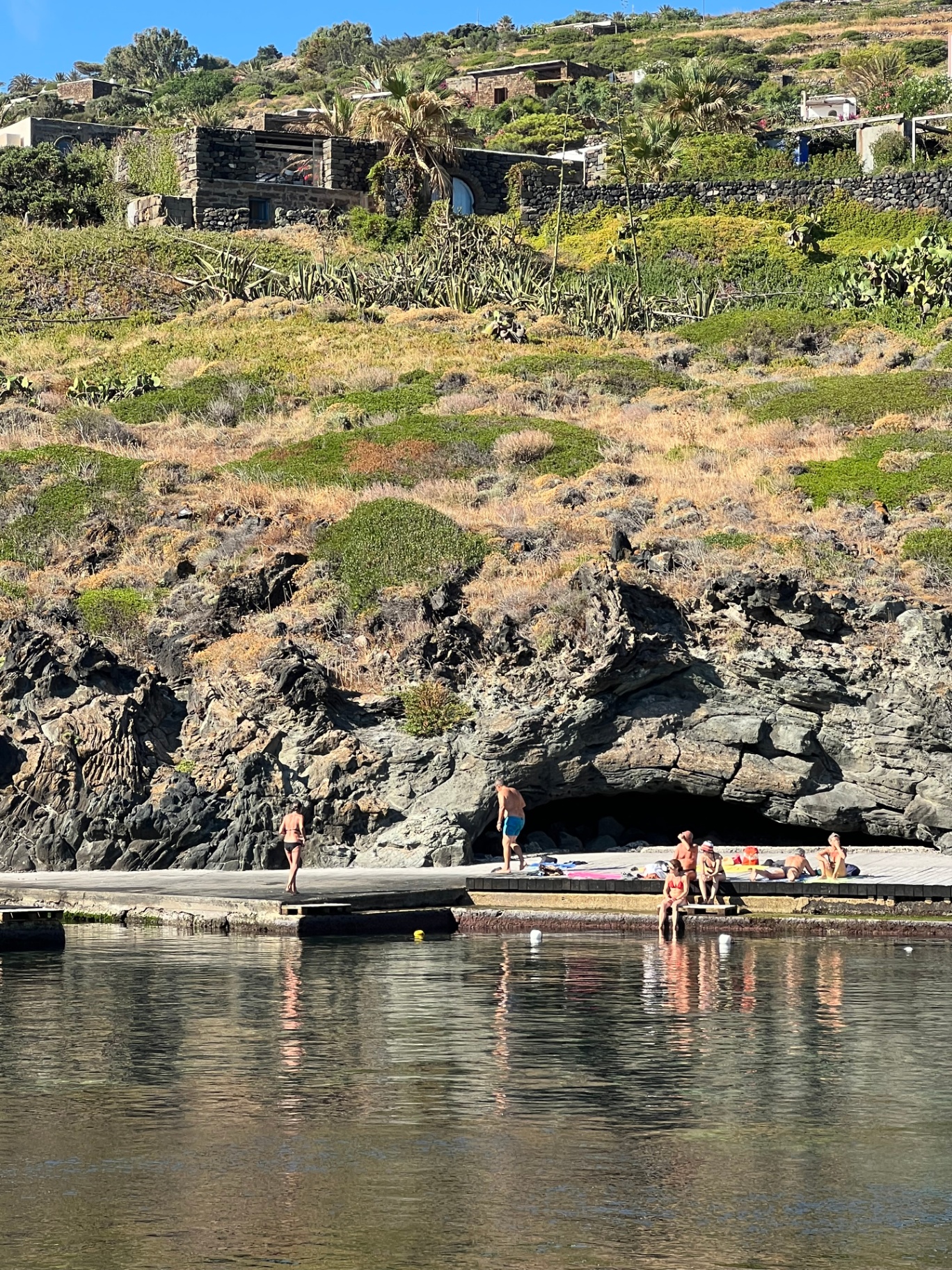 View from the bay below showing the stone house perched on the terraced hillside above the rocky coastline and swimming dock