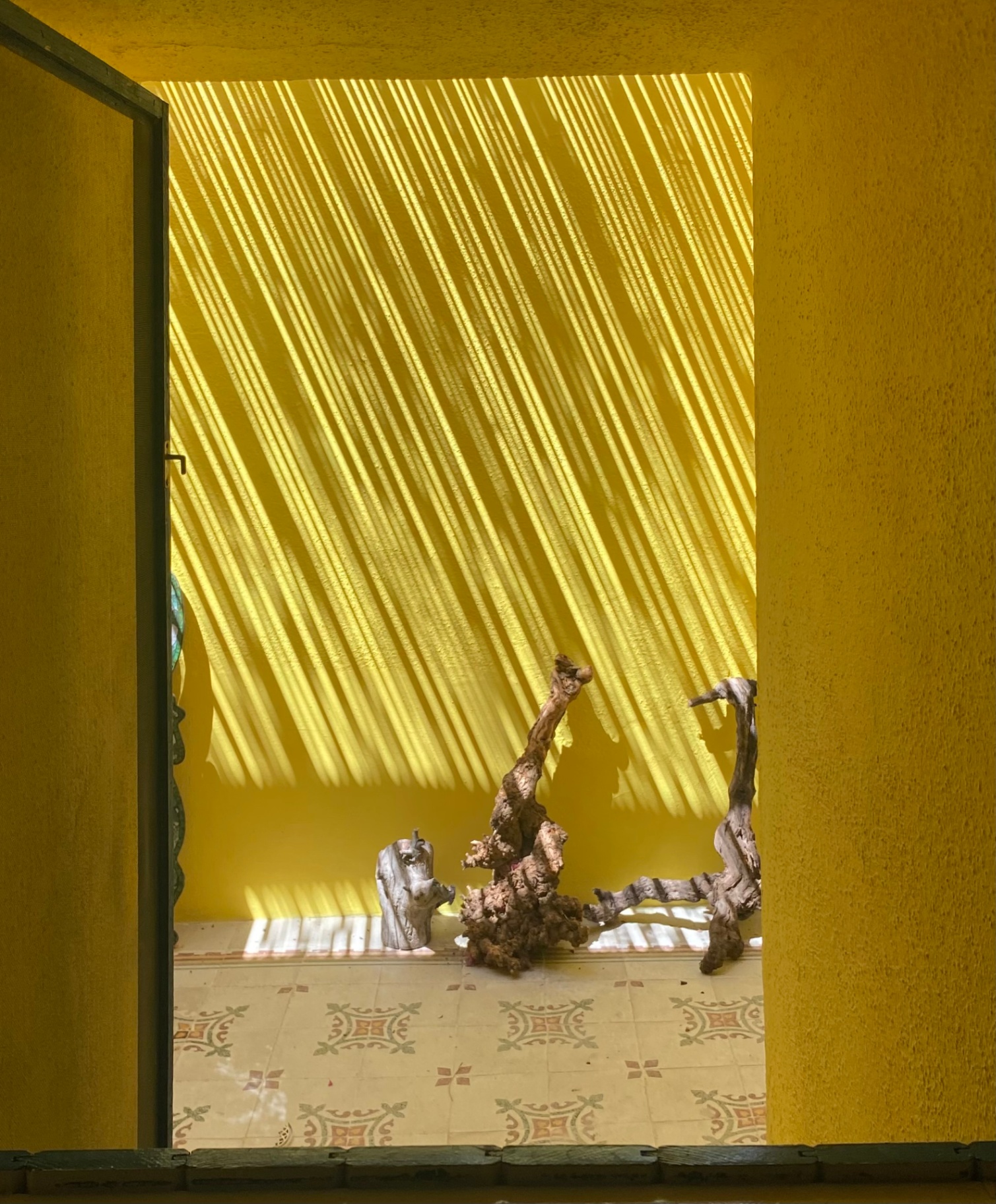 Yellow patio with striped shade casting diagonal light, driftwood sculptures, and patterned tile floor